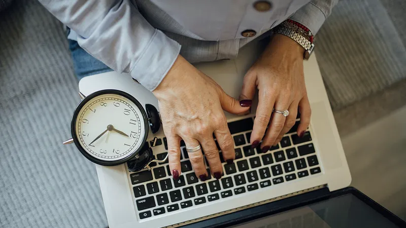 Hands typing on a laptop next to an alarm clock